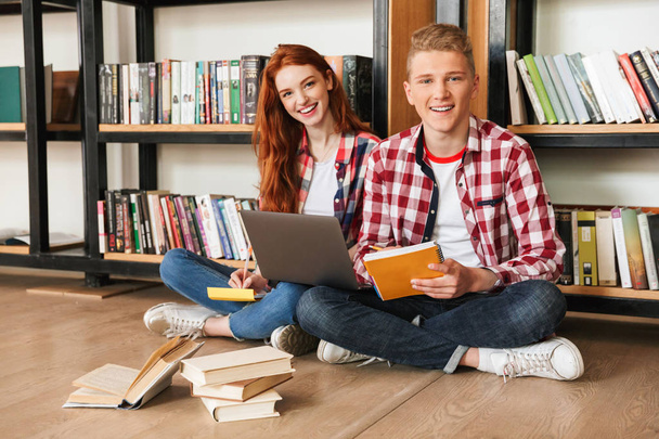 Happy teenage couple sitting on a floor at the bookshelf in a library and doing homework with laptop computer - Фото, изображение