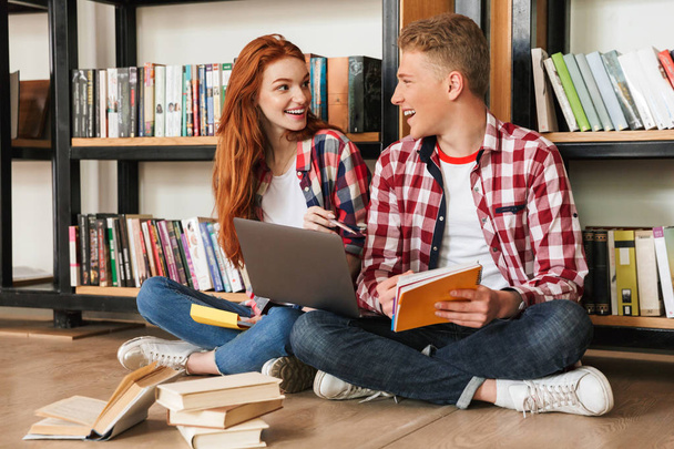 Cheerful teenage couple sitting on a floor at the bookshelf in a library and doing homework with laptop computer - Foto, Imagen