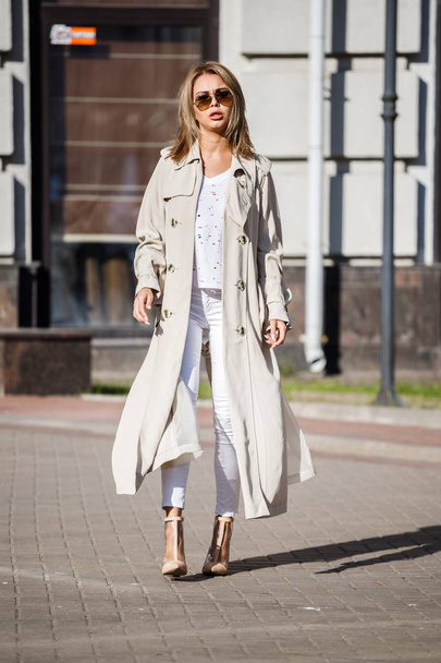 Outdoor portrait of beauty blond woman wearing beige jacket, white t-shirt and white pants, standing on street with blurred building on background - Fotó, kép