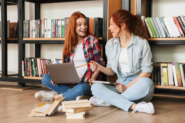 Two excited teenage girls sitting on a floor at the bookshelf in a library and doing homework with laptop computer - Foto, Imagen