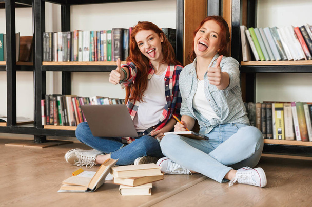 Two confident teenage girls sitting on a floor at the bookshelf in a library and doing homework with laptop computer - Foto, Imagem