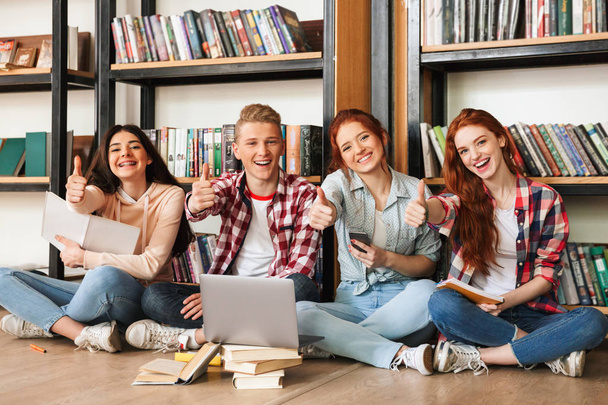 Group of confident teenagers doing homework while sitting on a floor at the library with laptop computer and mobile phone - Фото, зображення