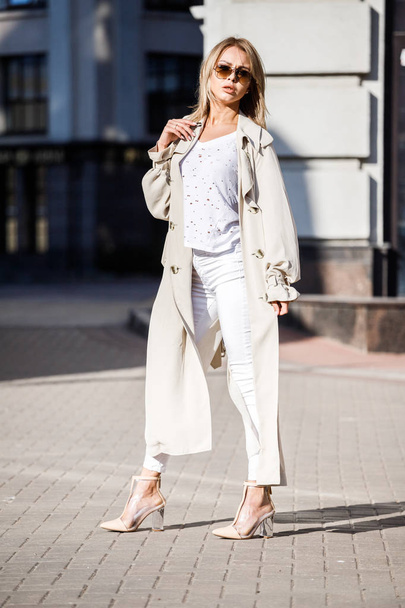 Outdoor Portret van schoonheid blonde vrouw beige jas, wit t-shirt en witte broek dragen, permanent op straat met wazig gebouw op achtergrond - Foto, afbeelding