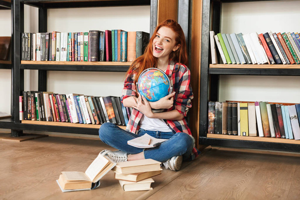 Image of happy young redhead pretty lady sitting in library with books and globe looking camera. - Фото, зображення