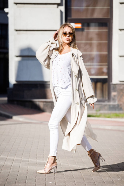 Outdoor portrait of beauty blond woman wearing beige jacket, white t-shirt and white pants, standing on street with blurred building on background - Foto, Imagem