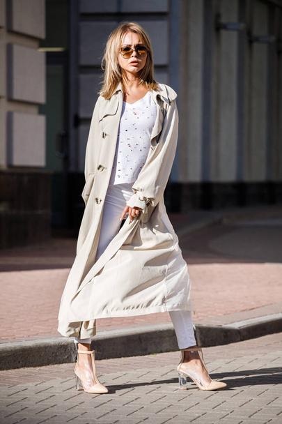 Outdoor portrait of beauty blond woman wearing beige jacket, white t-shirt and white pants, standing on street with blurred building on background - Zdjęcie, obraz