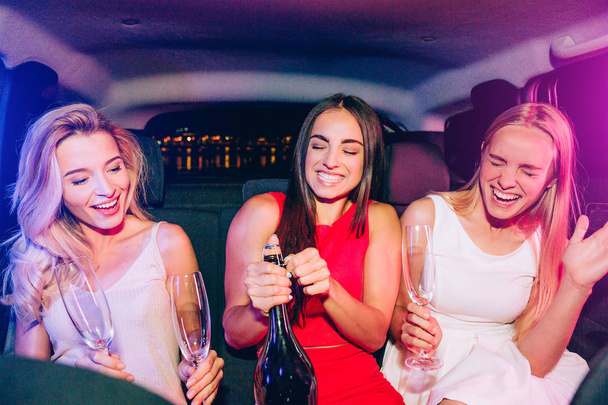 Beautiful girls are sitting in car. Brunette is opening bottle with alcohol. Blonde girls are very excited about that. Young women are holding glasses for champagne. - Photo, Image