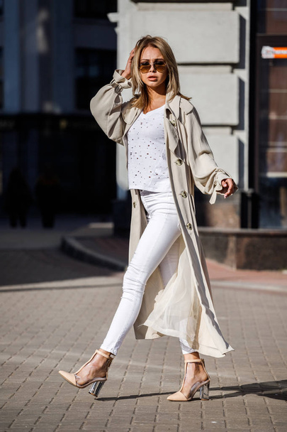Outdoor portrait of beauty blond woman wearing beige jacket, white t-shirt and white pants, standing on street with blurred building on background - Fotoğraf, Görsel
