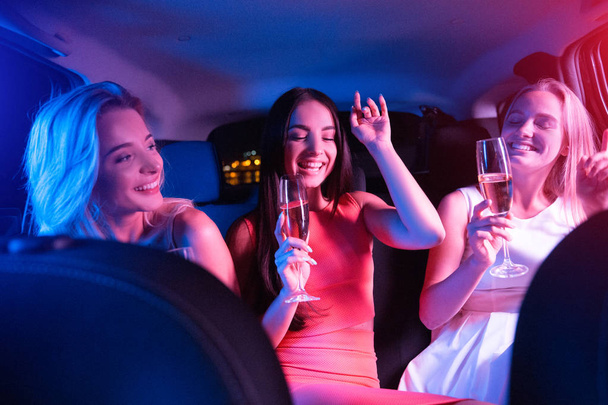 Colorful picture of three girls hanging out together. They are waving hands and enjoying the moment. Girls are holding glasses with champagne. They are sitting in car. - Photo, Image