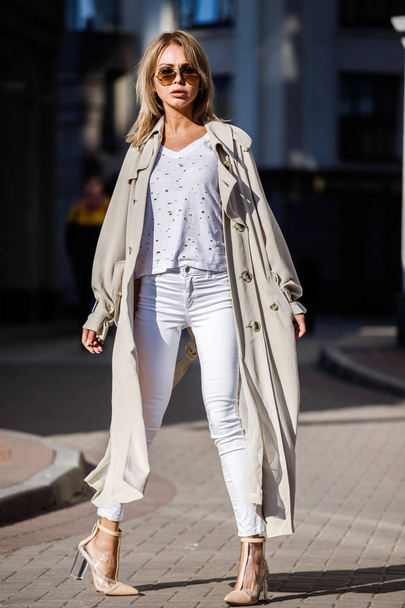 Outdoor portrait of beauty blond woman wearing beige jacket, white t-shirt and white pants, standing on street with blurred building on background - Fotografie, Obrázek