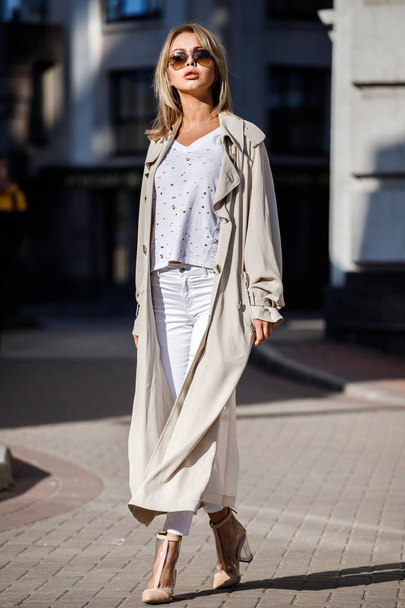 Outdoor portrait of beauty blond woman wearing beige jacket, white t-shirt and white pants, standing on street with blurred building on background - 写真・画像