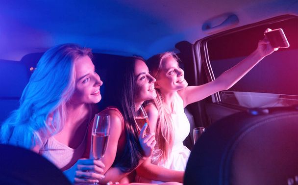 Menina loira está sentada no carro com seus amigos e tomar sefie. Eles estão segurando copos com champanhe e sorrindo na câmera. Meninas estão se divertindo
. - Foto, Imagem