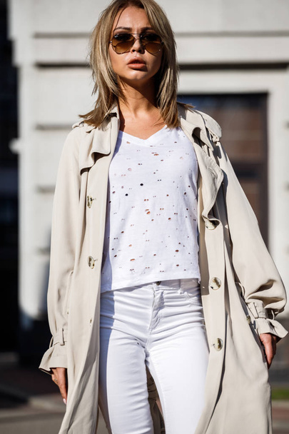 Outdoor portrait of beauty blond woman wearing beige jacket, white t-shirt and white pants, standing on street with blurred building on background - Foto, Bild