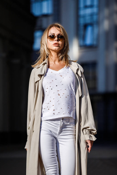 Outdoor portrait of beauty blond woman wearing beige jacket, white t-shirt and white pants, standing on street with blurred building on background - Fotografie, Obrázek