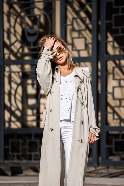 Outdoor portrait of beauty blond woman wearing beige jacket, white t-shirt and white pants, standing on street with blurred building on background - Foto, afbeelding