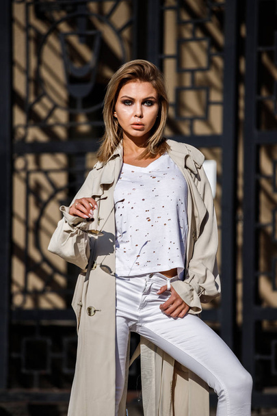 Outdoor portrait of beauty blond woman wearing beige jacket, white t-shirt and white pants, standing on street with blurred building on background - Foto, Imagen