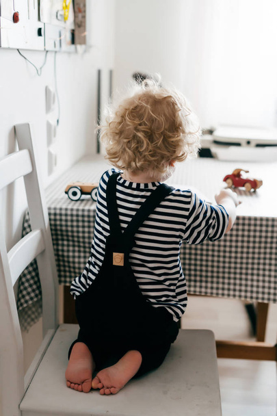 small curly baby boy in a striped blouse sits on a chair in the kitchen and draws in a notebook - Foto, afbeelding