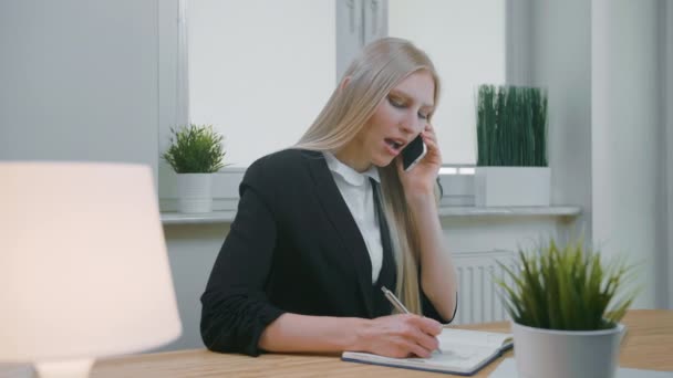 オフィスでスマートフォンで話しているビジネス女性。職場に座って携帯電話で交渉するオフィススーツ姿のエレガントな若いブロンドの女性ノートブックに必要な情報を手書きで - 映像、動画