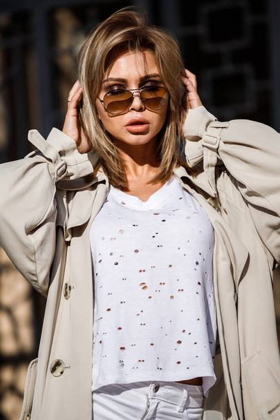 Outdoor portrait of beauty blond woman wearing beige jacket, white t-shirt and white pants, standing on street with blurred building on background - Photo, Image