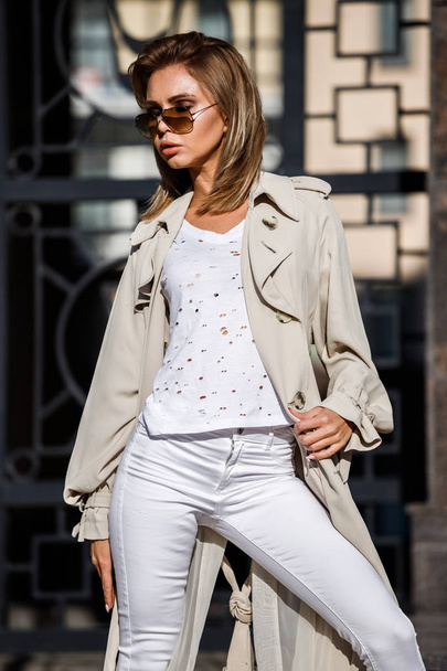 Outdoor portrait of beauty blond woman wearing beige jacket, white t-shirt and white pants, standing on street with blurred building on background - Foto, imagen