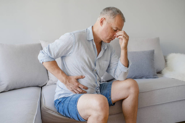 Terrible stomachache. Frustrated handsome young man hugging his belly and keeping eyes closed. Businessman stomachache. Mature man sitting with stomach pain - Photo, Image