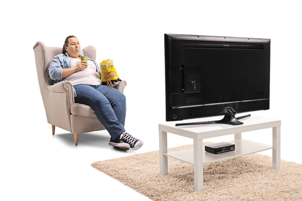 Overweight woman with a bag of chips and a drink sitting in an armchair and watching television isolated on white background - Fotó, kép