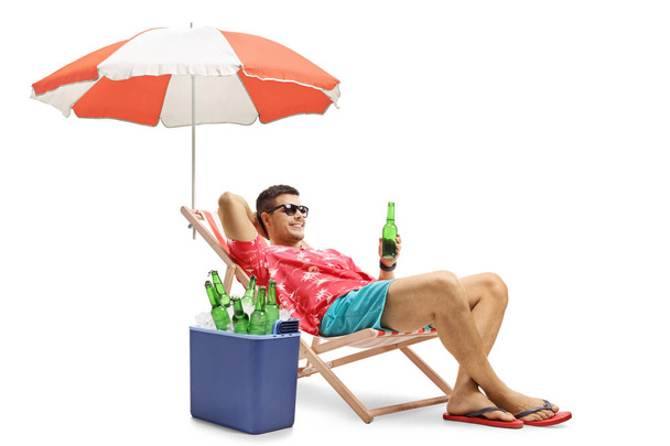Tourist with a bottle of beer relaxing in a deck chair with an umbrella next to a cooling box isolated on white background - Фото, зображення