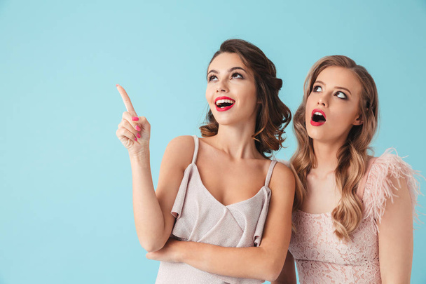 Two beautiful girls 20s wearing party outfit pointing index fingers upward at copyspace with excited faces isolated over blue background - Photo, image