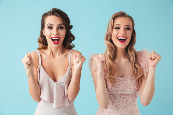 Two Surprised happy women in dresses screaming and rejoices while looking at the camera over turquoise background - 写真・画像