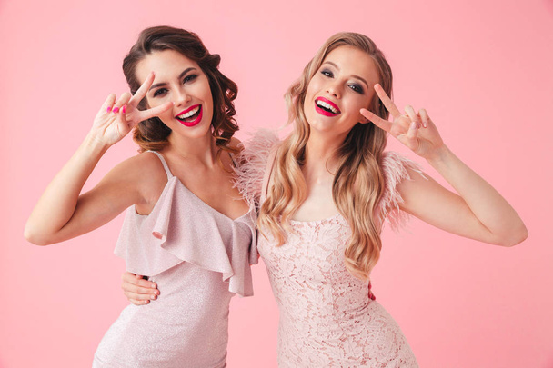 Two funny women in dresses having fun together while showing peace gestures and looking at the camera over pink background - Zdjęcie, obraz