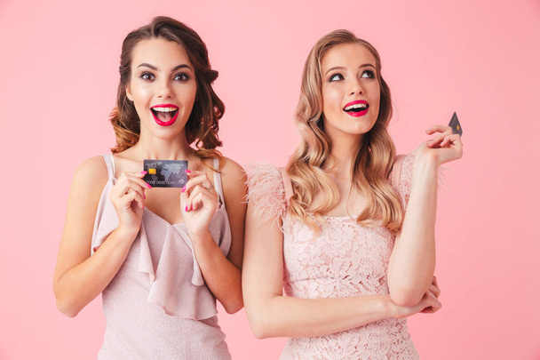 Two intrigued women in dresses posing with credit cards and looking around over pink background - Foto, immagini