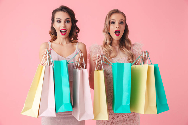 Two surprised happy women in dresses opening packages and looking at the camera over pink background - Photo, image