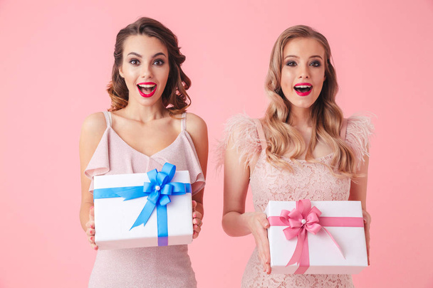 Two surprised happy women in dresses holding gift boxes and looking at the camera over pink background - Photo, Image