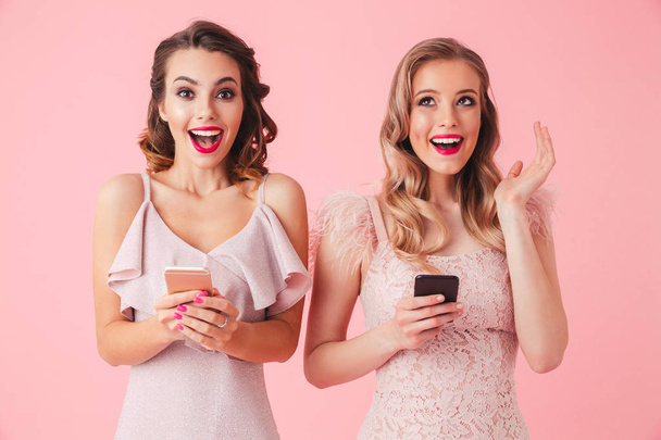 Two surprised happy elegant women in dresses posing together and holding their smartphones over pink background - 写真・画像