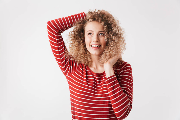 Playful blonde curly woman in casual clothes posing and looking away while touching her hair over grey background - Fotografie, Obrázek