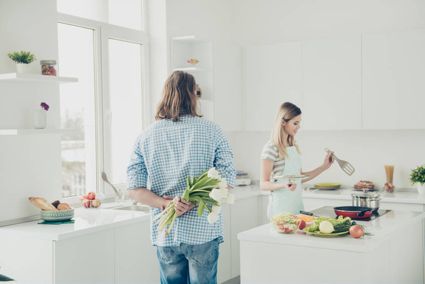 Portrait of handsome man holding bouquet of white tulips behind back in hands preparing surprise while housewife making meal dinner in modern kitchen with interior - Foto, immagini