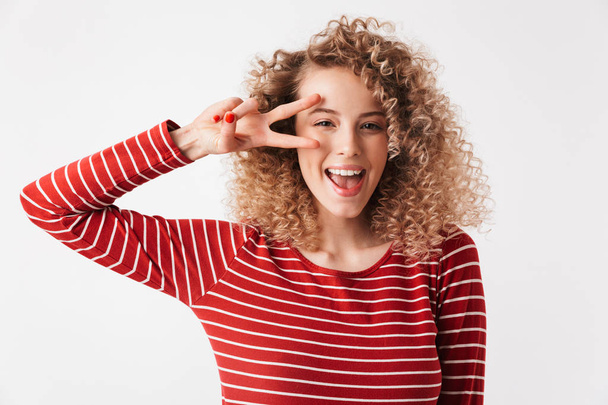 Cheerful happy blonde curly woman in casual clothes showing peace gesture and looking at the camera over grey background - Fotoğraf, Görsel
