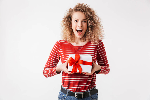 Portrait of happy young girl with curly hair holding present box isolated over white background - Фото, изображение
