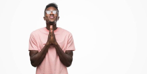 Joven afroamericano vestido con camiseta rosa rezando con las manos juntas pidiendo perdón sonriendo confiado
. - Foto, Imagen