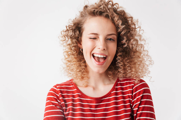 Cheerful blonde curly woman in casual clothes having fun and winks while looking at the camera over grey background - Photo, image