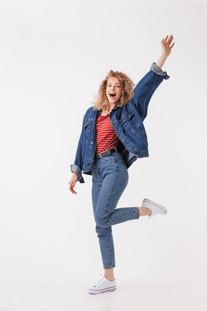 Full length image of Cheerful blonde curly woman in denim clothes rejoices and looking at the camera over grey background - Fotó, kép