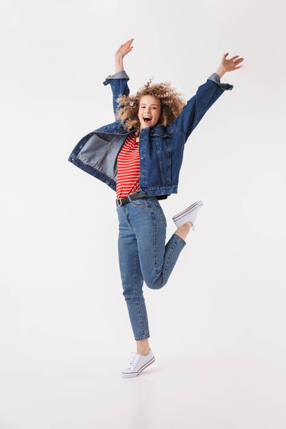 Full length image of Happy blonde curly woman in denim clothes jumping and rejoices while looking at the camera over grey background - Fotó, kép