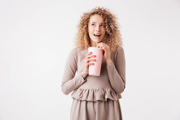Portrait of a cheerful young curly blonde girl in dress drinking from a plastic cup isolated over white background - Foto, Imagem