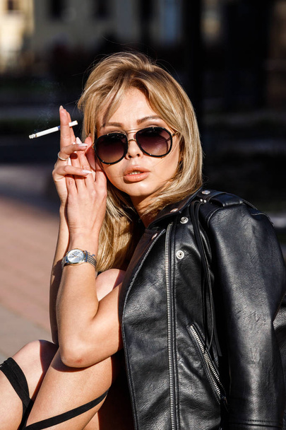 Outdoor portrait of beauty blond woman wearing black leather jacket and black shoes on high heels, sitting on border and smoking cigarette  - Fotografie, Obrázek