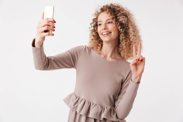 Portrait of a pretty young curly blonde girl in dress taking selfie with mobile phone isolated over white background - Fotoğraf, Görsel