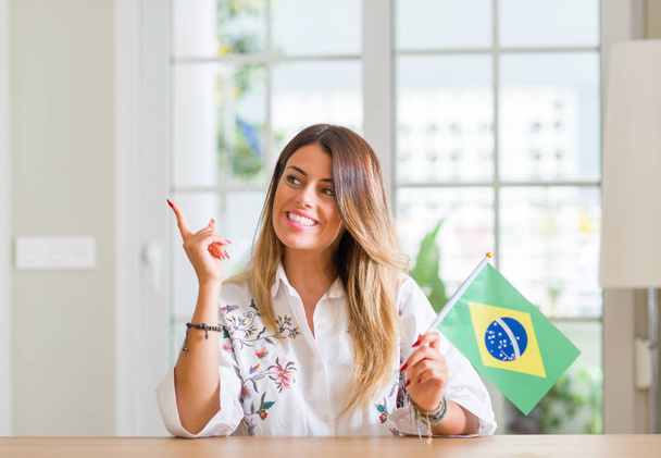 Jovem mulher em casa segurando bandeira do Brasil muito feliz apontando com a mão e o dedo para o lado
 - Foto, Imagem