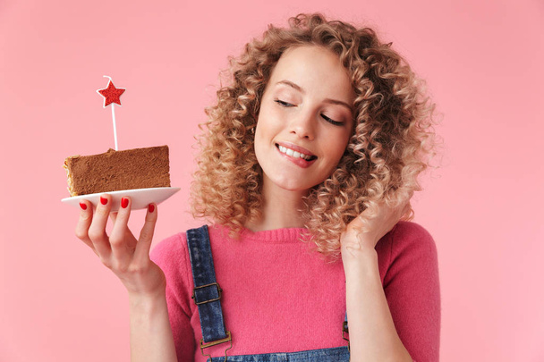Portrait of hungry young girl with curly hair looking at cupcake on a plate isolated over pink background - 写真・画像
