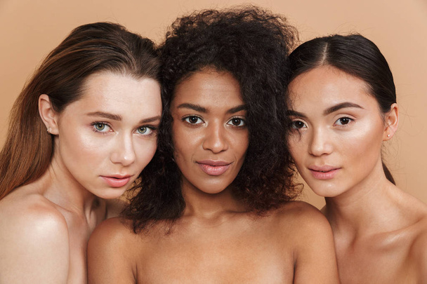 Close up image of three sensual naked women posing together and looking at the camera over beige background - Zdjęcie, obraz
