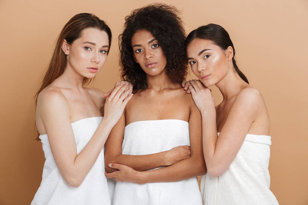 Three attractive women wearing in towels posing together and looking at the camera over beige background - Photo, image