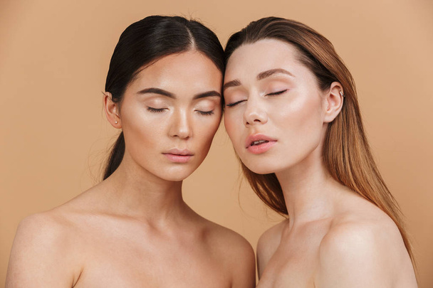 Beauty portrait closeup of two different nation women, asian and caucasian nude girls with closed eyes posing together at camera isolated over beige background - Photo, Image